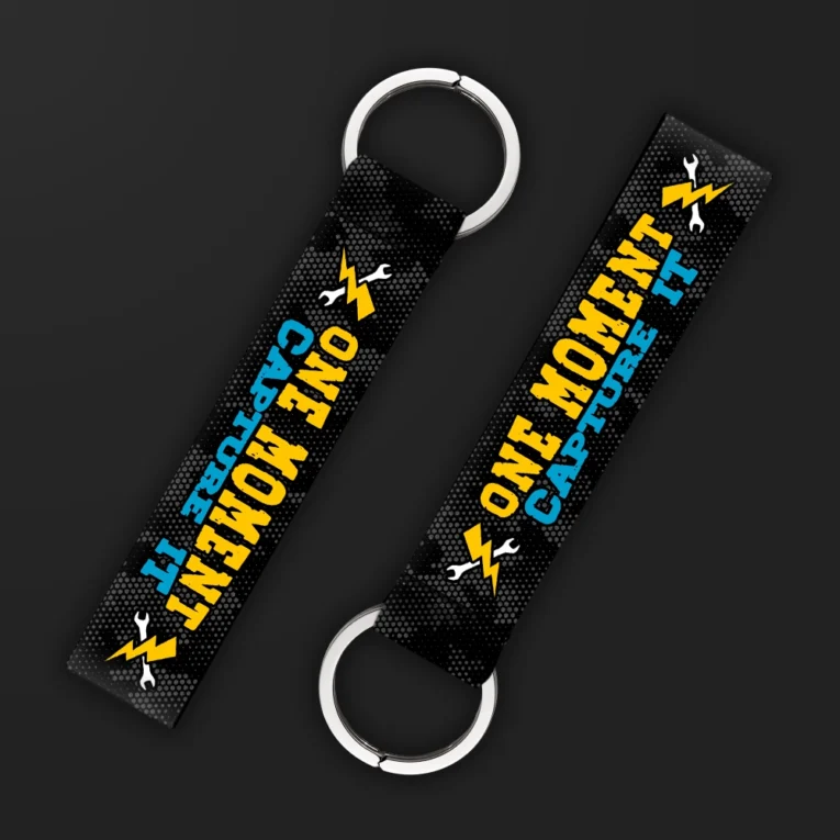 Inline-4 One Moment keychain strap with yellow and blue 'ONE MOMENT CAPTURE IT' text and wrench-and-lightning icons on black, double keyring