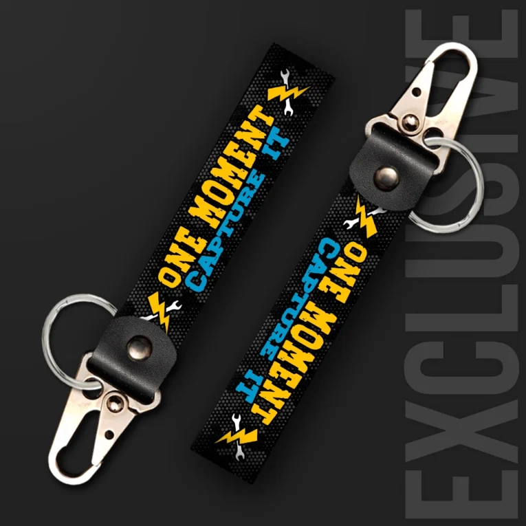 Inline-4 One Moment Capture It keychain strap with yellow and blue 'ONE MOMENT CAPTURE IT' text and wrench-and-lightning icons on black, swivel hook clip + keyring