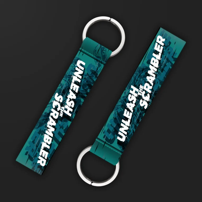 Inline-4 Unleash the Scrambler keychain strap with white 'UNLEASH THE SCRAMBLER' text over a teal off-road motorcycle illustration, double keyring