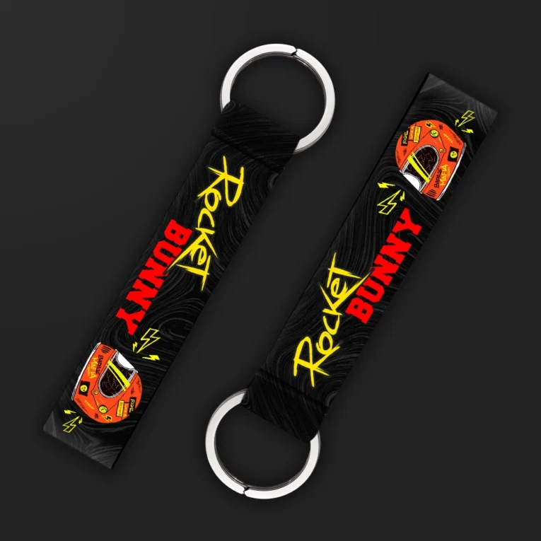 Inline-4 Rocket Bunny keychain strap with yellow 'Rocket Bunny' script, lightning icons, and a racing helmet graphic on black, double keyring
