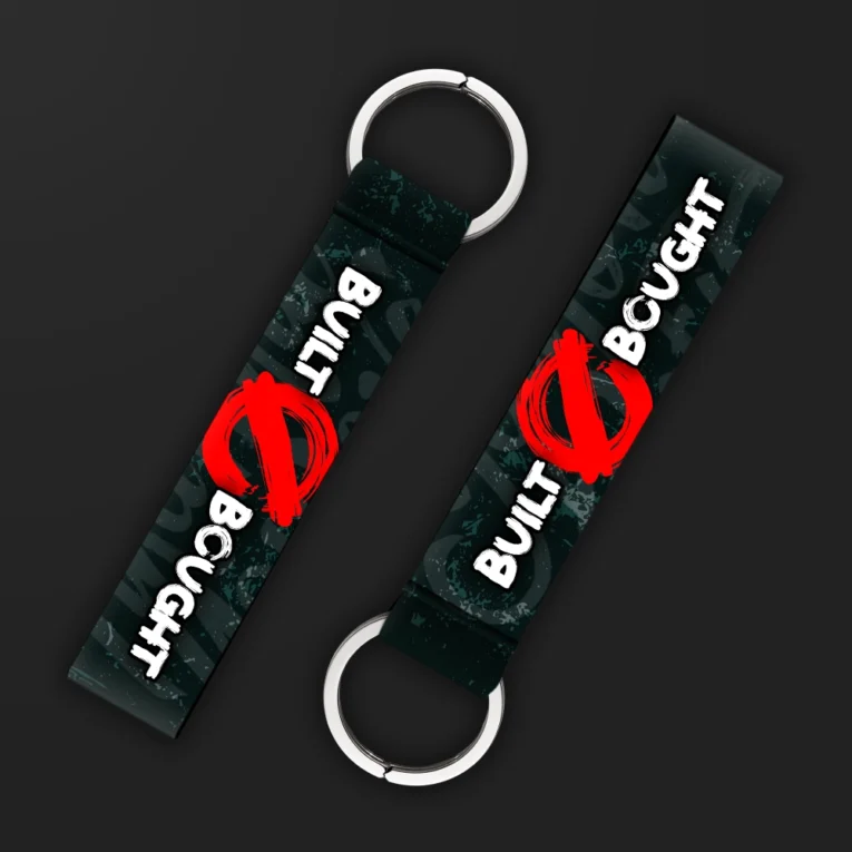 Inline-4 Built Not Bought keychain strap with white 'BUILT' and 'BOUGHT' text crossed out by a red brush stroke on a dark camo background, double keyring
