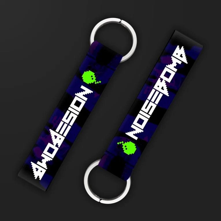 Inline-4 Noisebomb keychain strap with a retro pixel-art skull-bomb icon and 'NOISEBOMB' wordmark on a purple camo print, double keyring